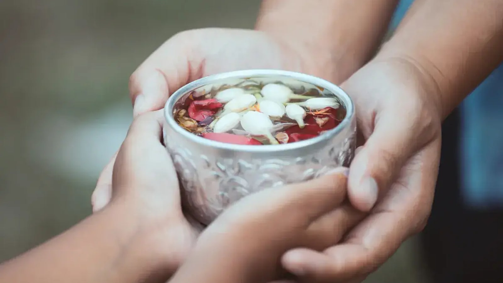 An adult's hands and a child's hands holding a small bowl of water together, flower petals floating on the surface